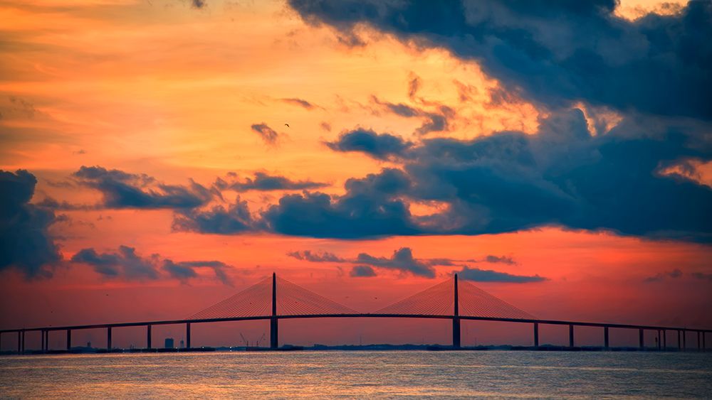 The Skyway Bridge over the Gulf of Mexico with the reds and oranges of the sunrise in the sky art print by Sheila Haddad for $57.95 CAD