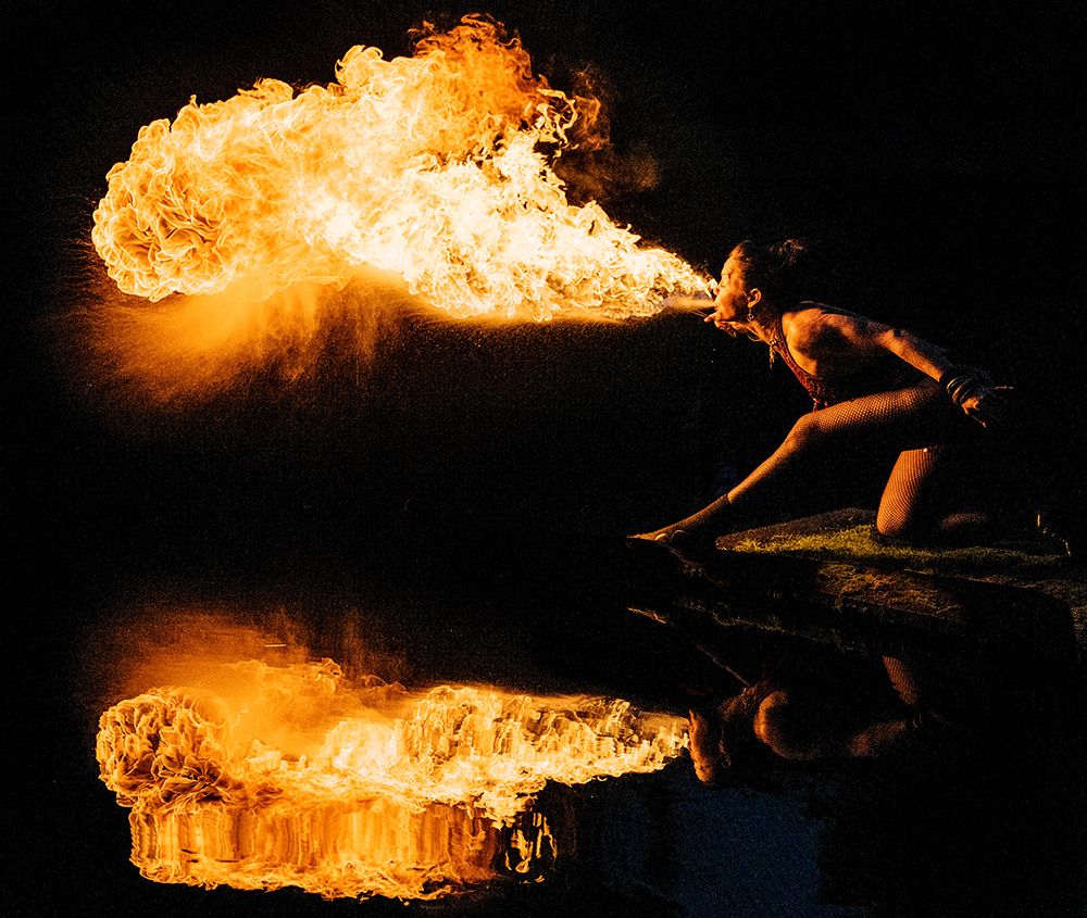 A Performer Breathes Fire, Creating A Vibrant Flame And Reflecting In Water Against A Dark Backdrop art print by Pete Saloutos for $57.95 CAD