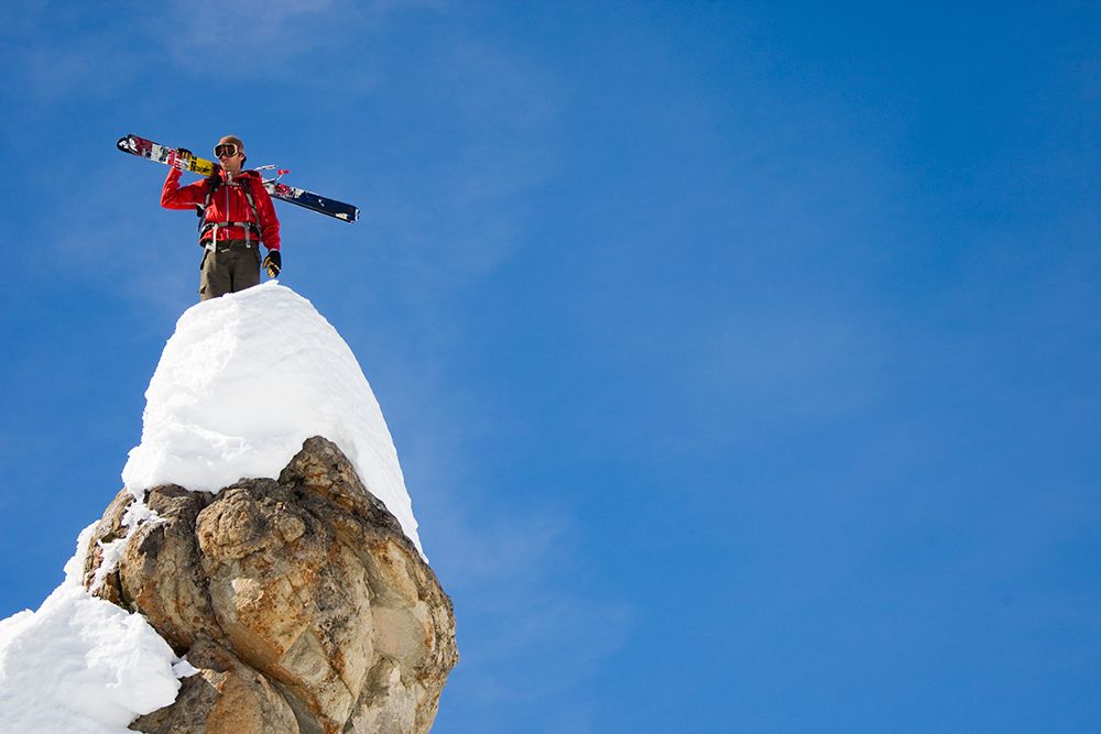Male Skier With Skis Over The Shoulder Perched On A Rock. Blue Sky Background. art print by Gabe Rogel for $57.95 CAD